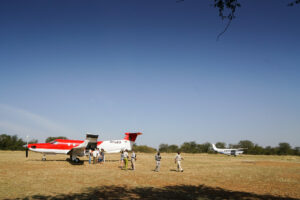 Single-engine bush aircraft parked on a remote dirt savannah airstrip preparing for passenger boarding.